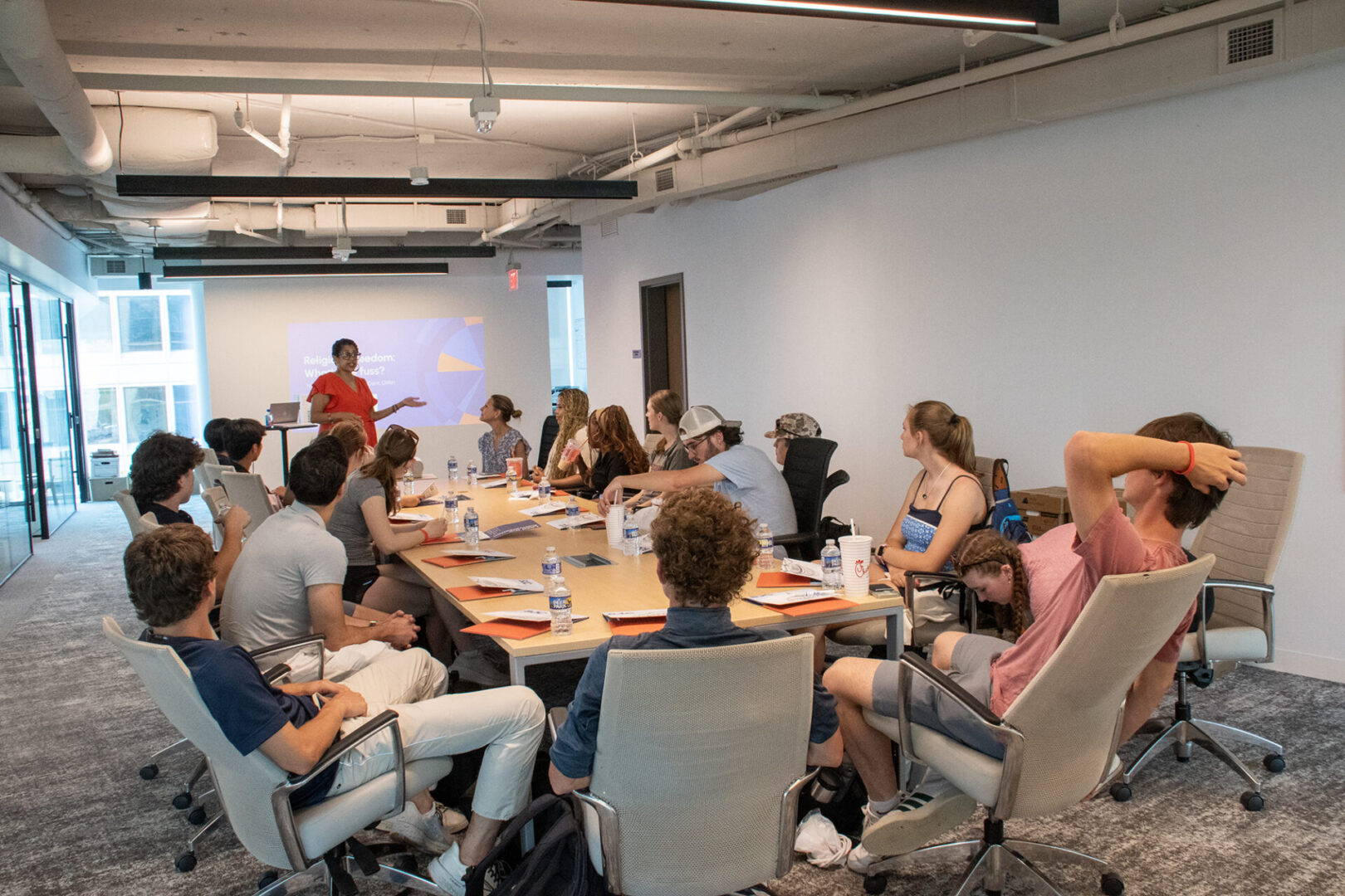 A group of people in a meeting room attentively listens to a presenter at the front.
