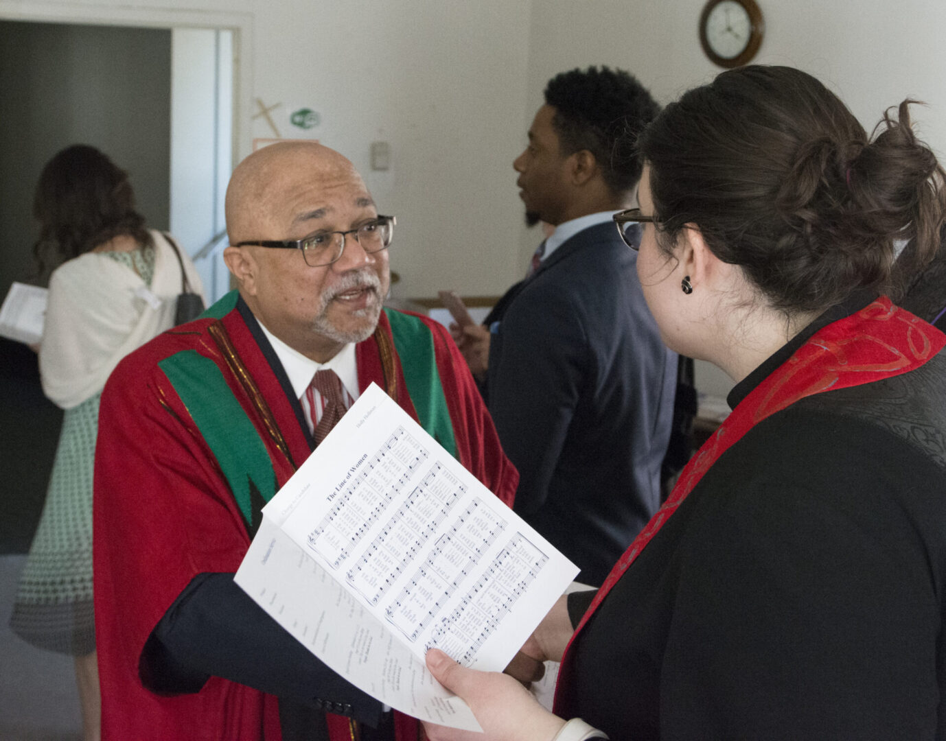 A man in a red robe is talking to a woman holding papers, with people in the background.