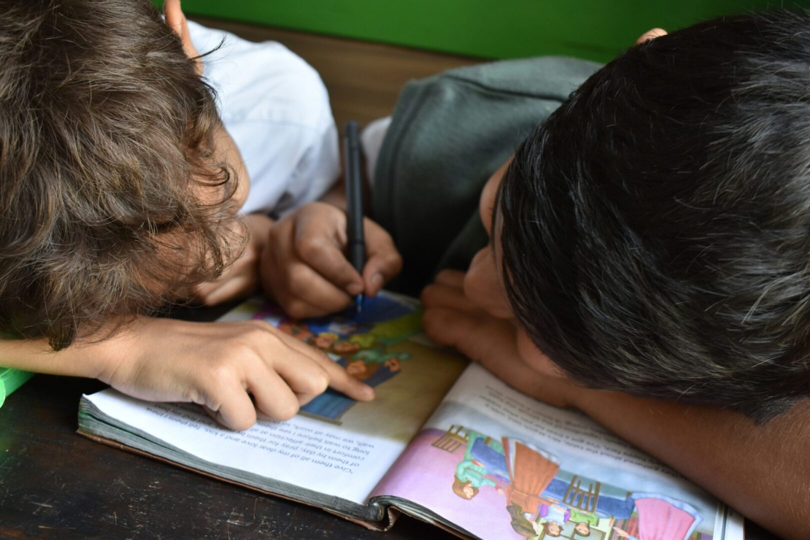 Two children are intently drawing and pointing at a colorful book.