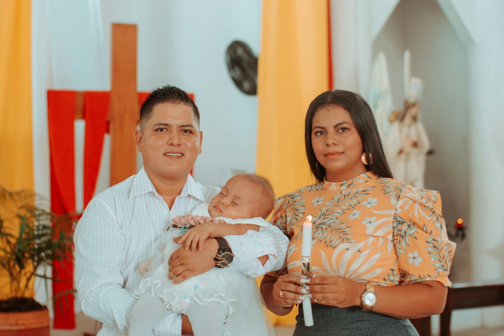 A couple stands together in a church, holding a baby and a candle, wearing formal attire.