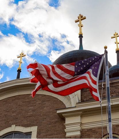 An American flag waves in front of a building with domes topped by crosses against a partly cloudy sky.