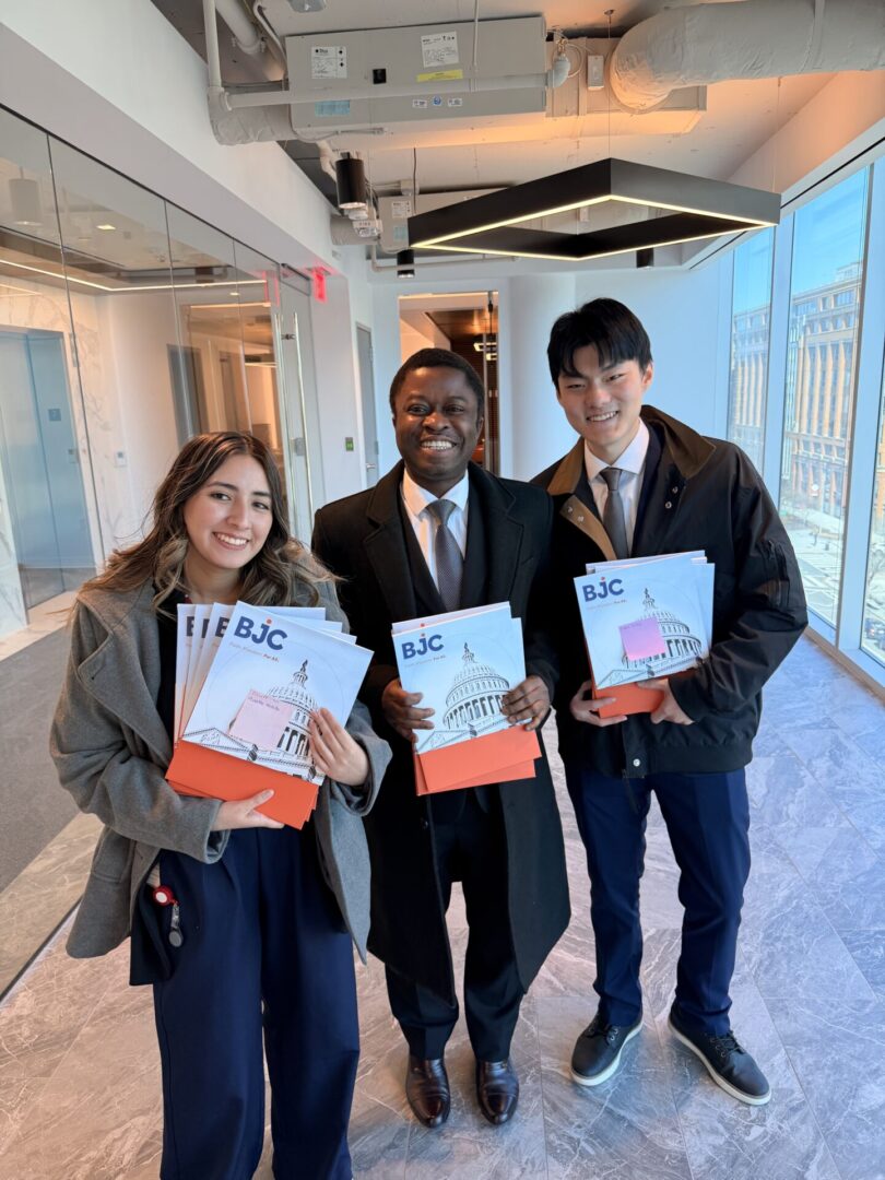 Three individuals stand indoors, smiling and holding documents in a modern office setting.