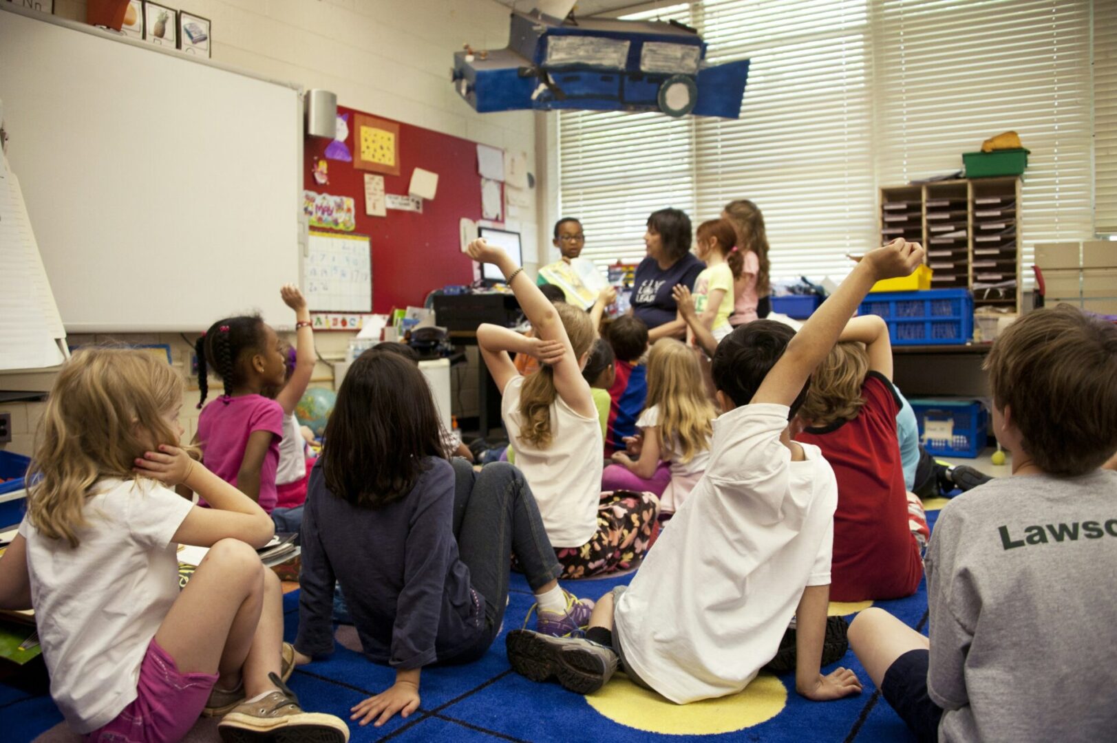 Students sit on the floor in a classroom, attentively raising their hands to participate.