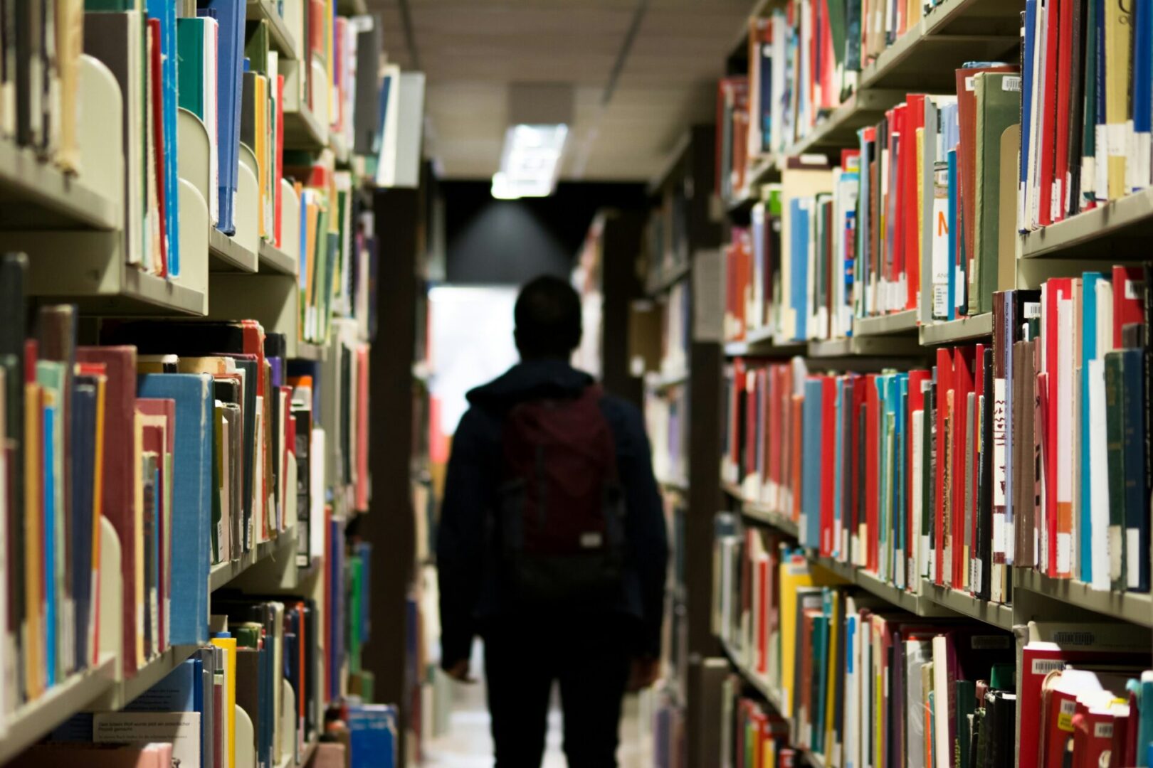 A person with a backpack walks through a library aisle lined with colorful books.