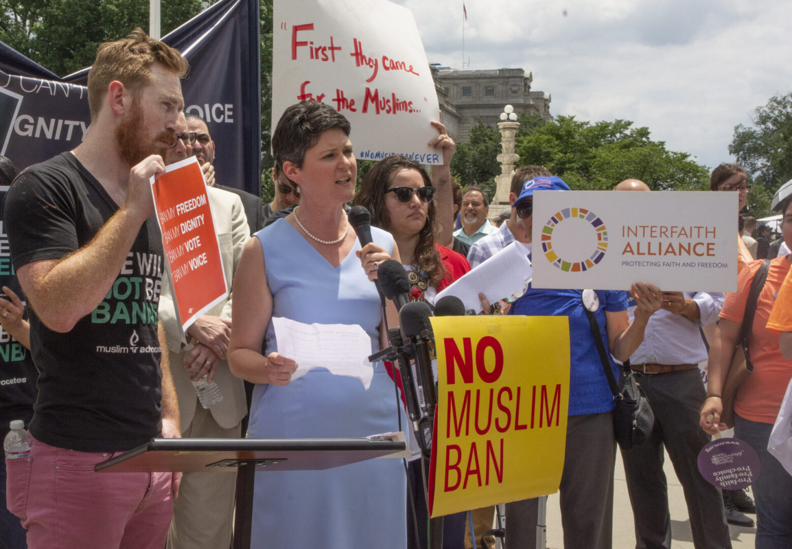 A group of people is gathered outdoors holding signs protesting against the Muslim ban.