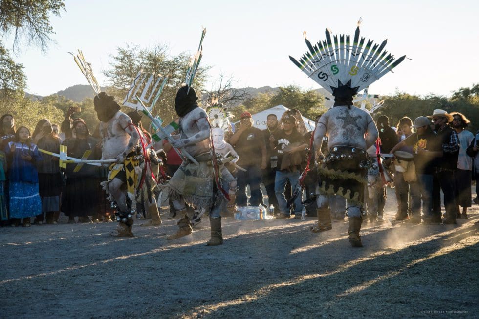 A group of people wearing traditional indigenous attire with feathered headdresses performs a ritual dance outdoors.