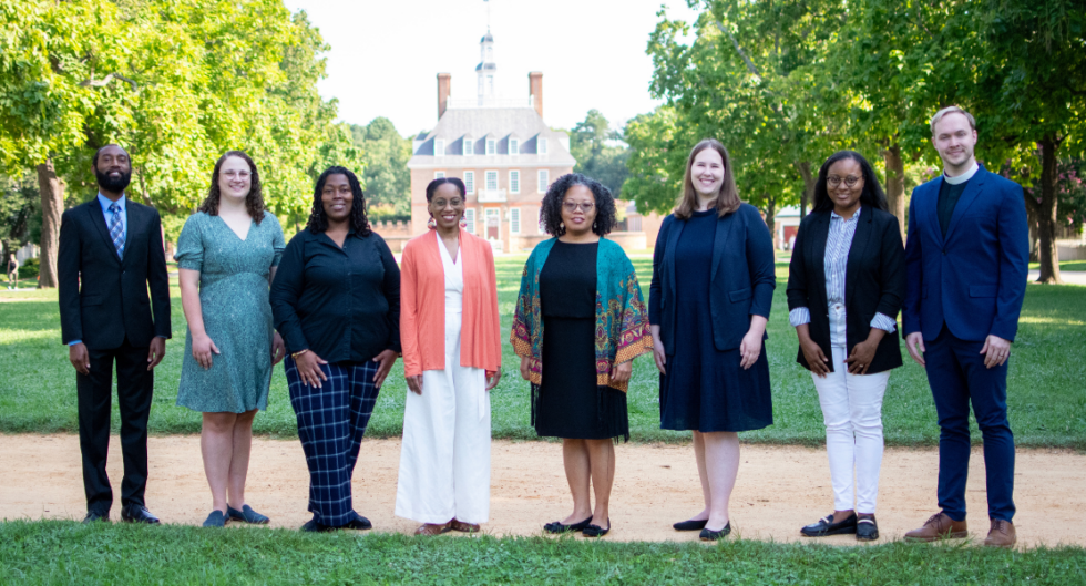 A group of individuals is standing on a pathway, with a historic building and lush greenery in the background.