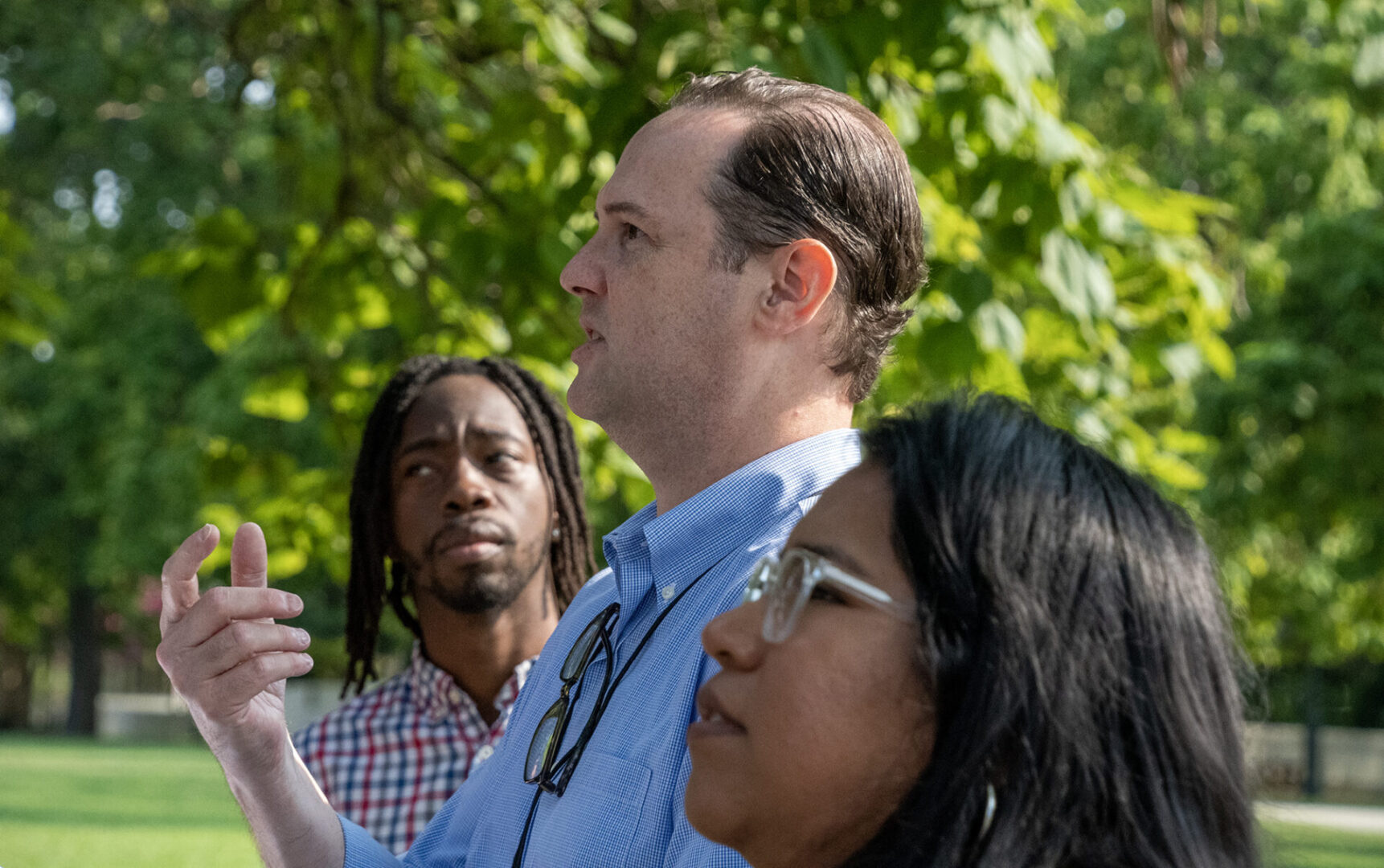 A man gestures while speaking outdoors, accompanied by two people listening attentively amidst a background of lush green trees.