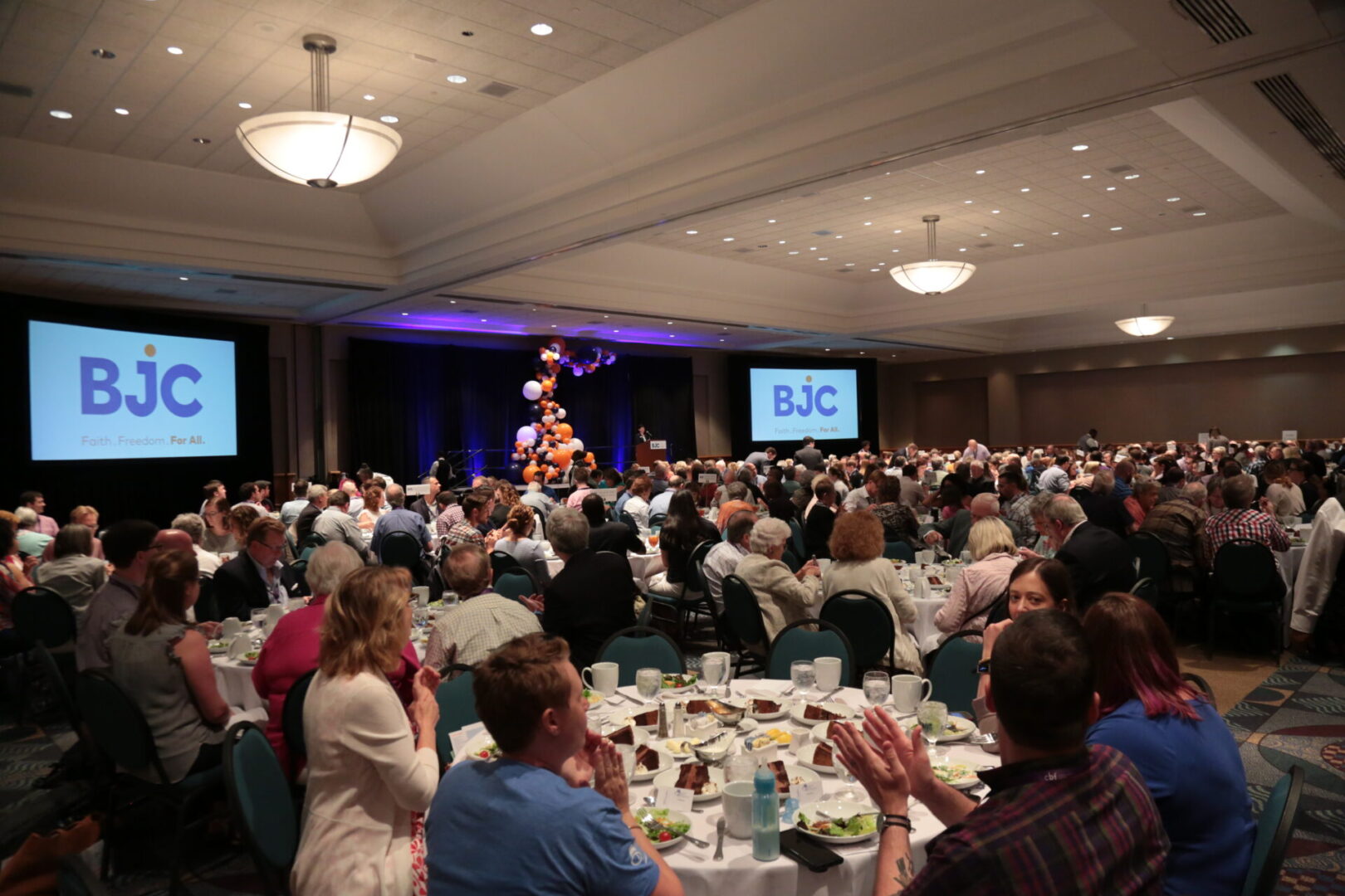 A large crowd is gathered in a banquet hall, watching a performer on stage with people seated at round tables.