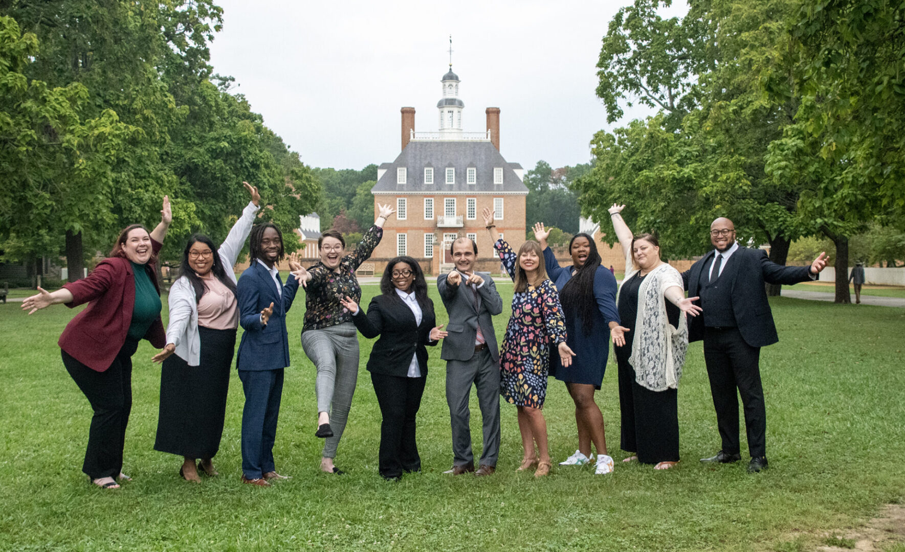 A group of people is standing outdoors, smiling and raising their hands, with a historic building in the background.