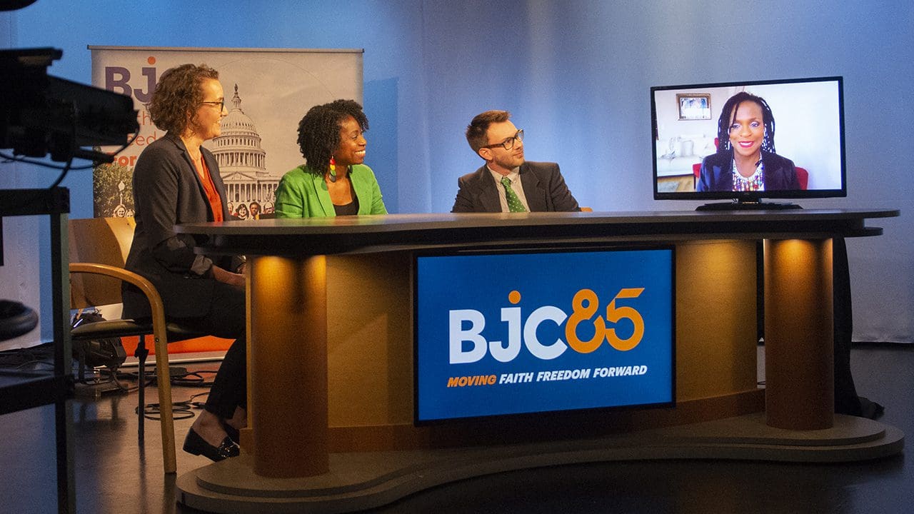 A panel of professionals is seated at a table, engaging in a discussion, with a BJC display and a TV screen featuring a video call participant.