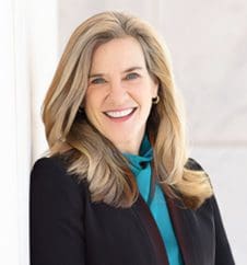 A person with long, light brown hair is smiling while wearing a black blazer and a teal blouse.