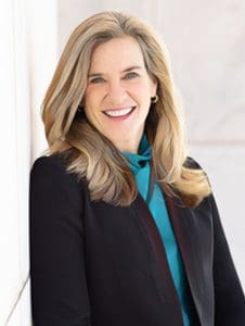 A person with long hair is smiling, dressed in a black blazer and turquoise top.