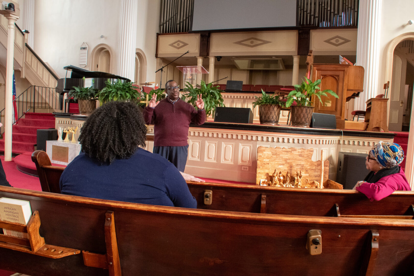 A person stands at the front of a church speaking, with plants and a piano in the background.