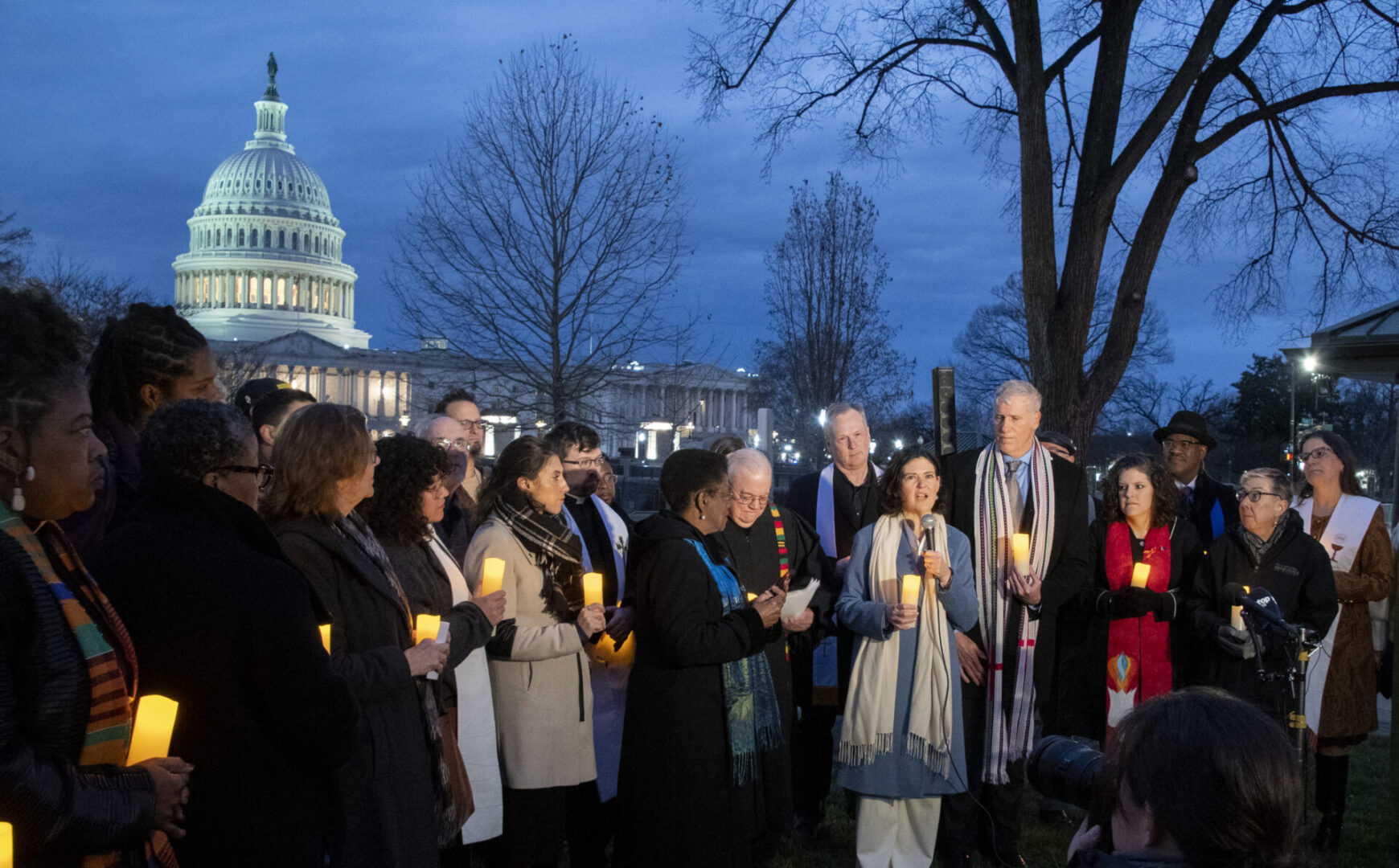 A group of people stands holding candles near a building with a dome, likely during an evening vigil.