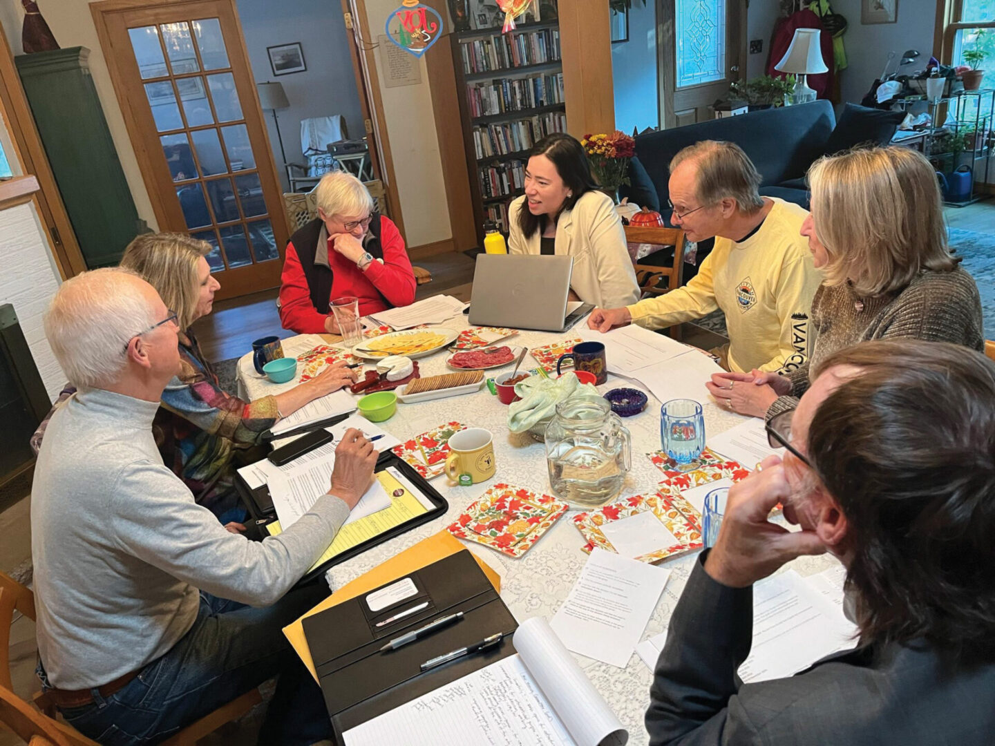A group of people is gathered around a table, engaged in discussion with notebooks and food items present.