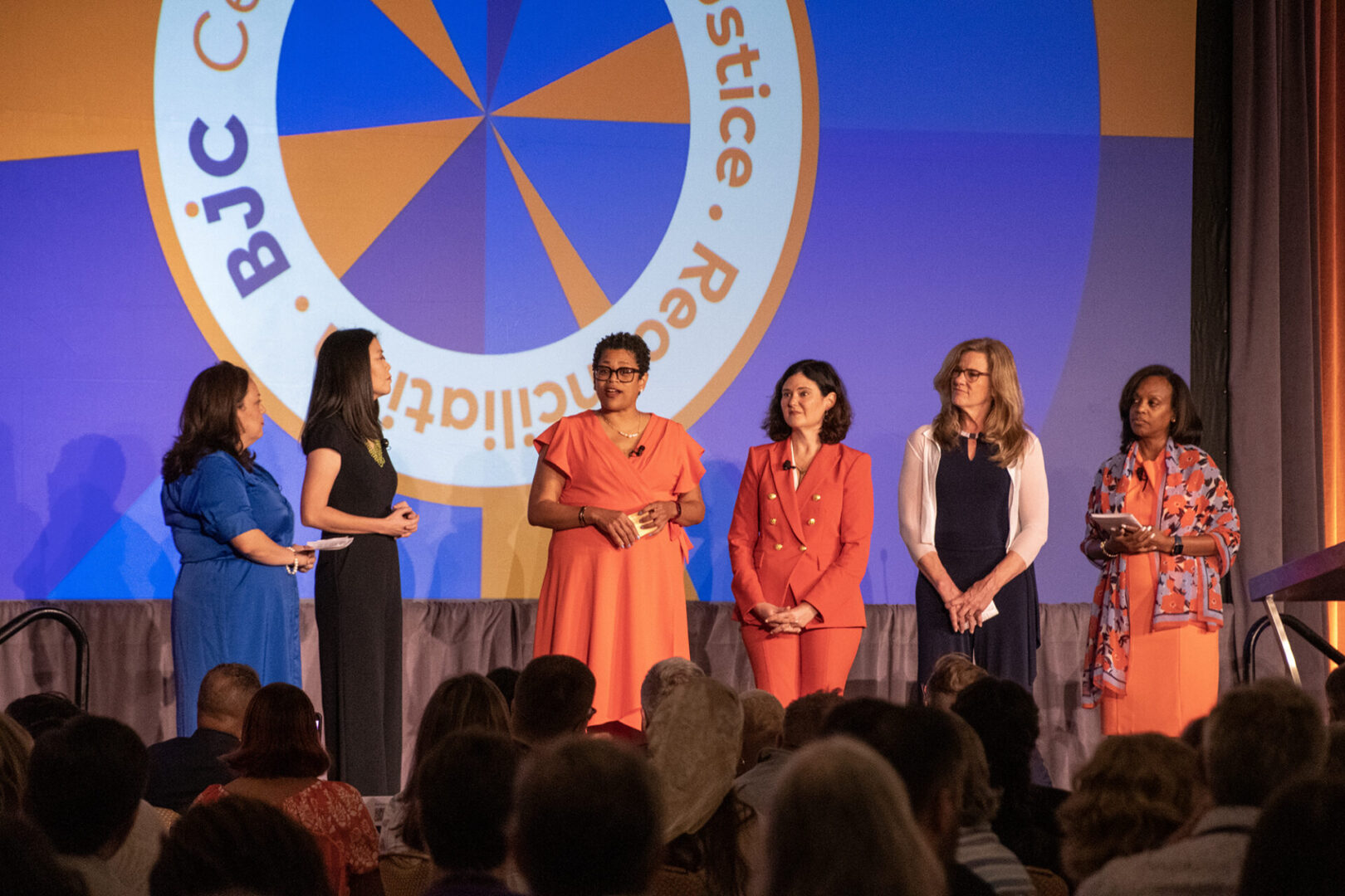 A group of people stands on stage in front of a colorful backdrop with text, engaging with an audience.