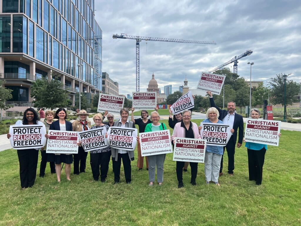 People are standing on grass holding signs with buildings and a crane in the background.