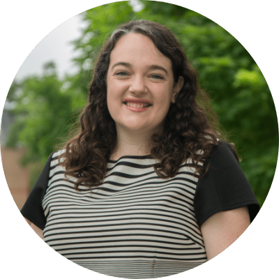 A person with curly hair is smiling outdoors, wearing a striped top with greenery in the background.