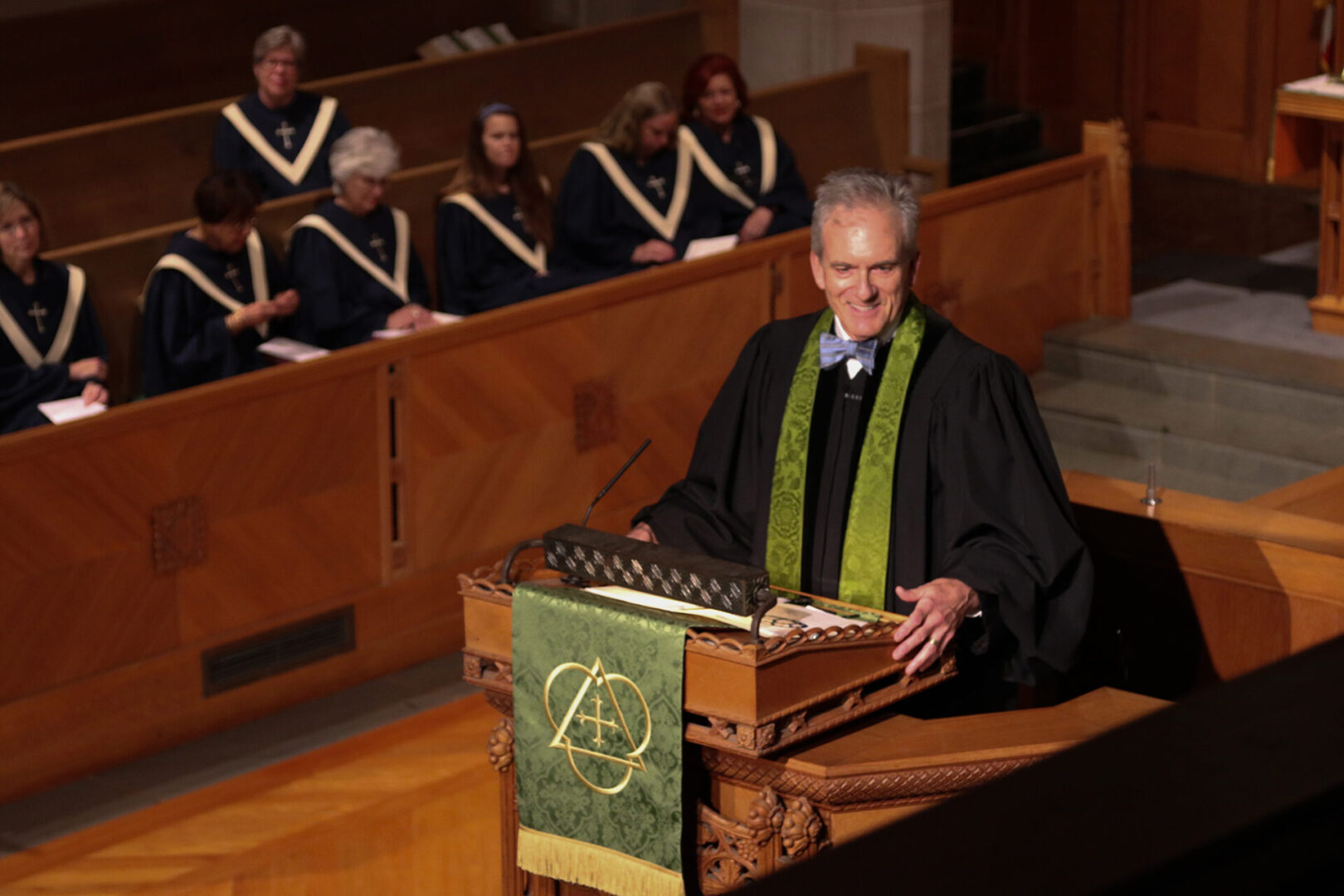 A person in a church setting stands at a lectern wearing a robe and green stole, with choir members seated behind.