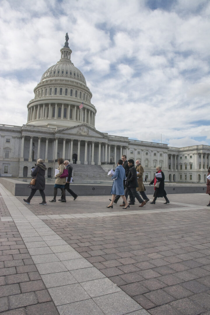A group of people walk past the United States Capitol building under a partly cloudy sky.