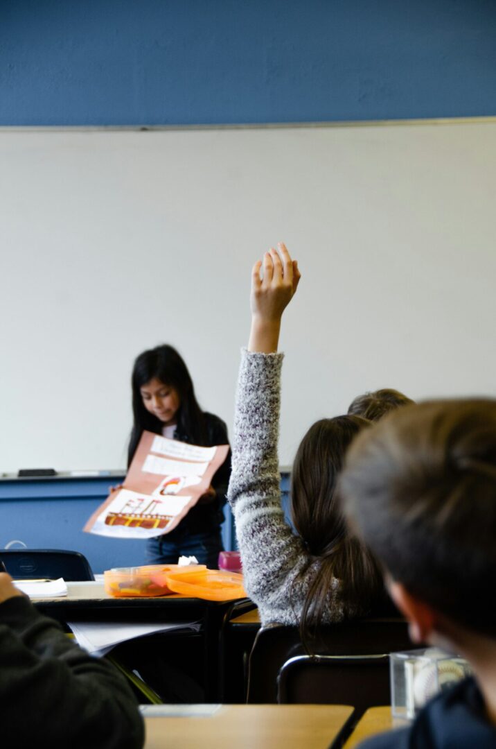 A person is raising their hand in a classroom while a teacher holds a book.