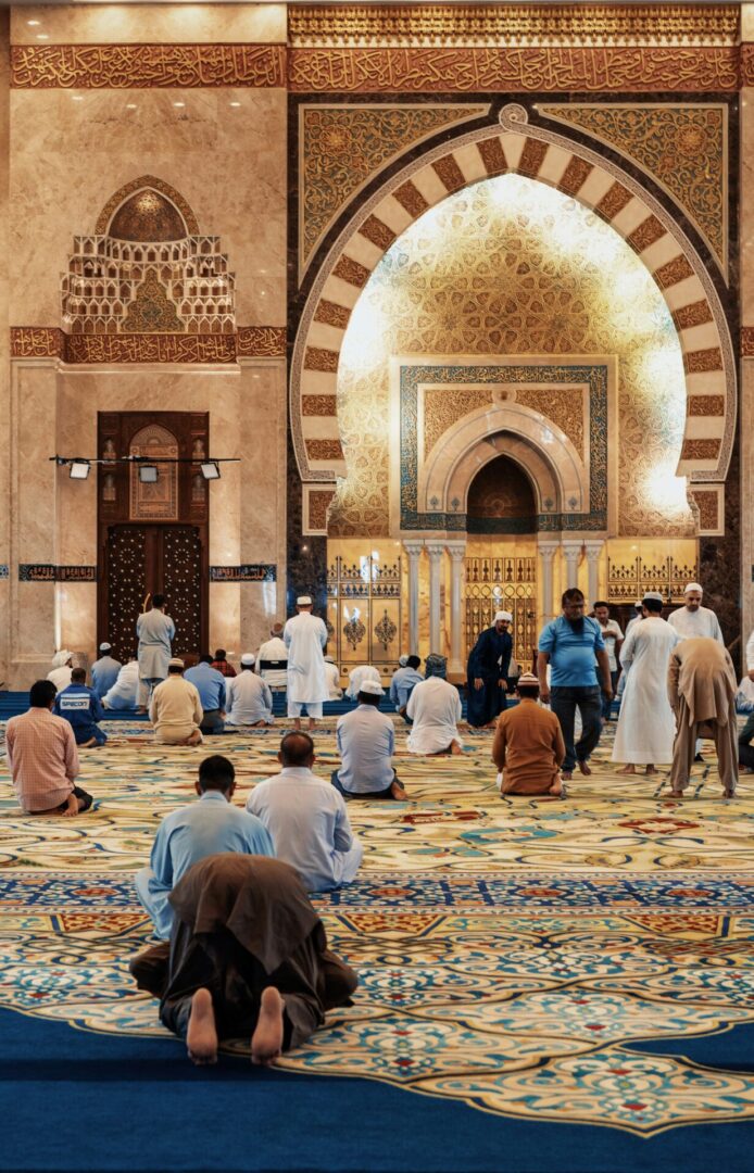 People are gathered inside a beautifully decorated mosque, praying on patterned carpets under ornate arches.