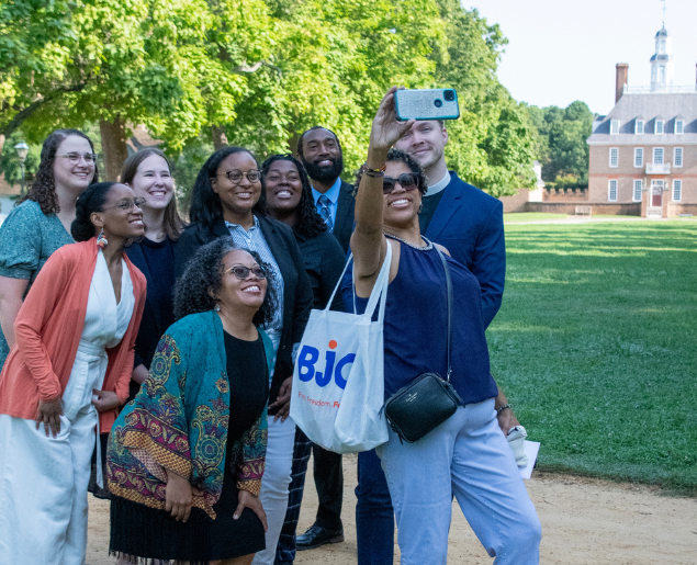 A group of people is smiling and taking a selfie outdoors on a sunny day.