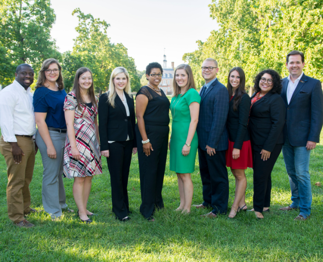 A diverse group of people is standing together outdoors on a grassy area, with trees in the background.