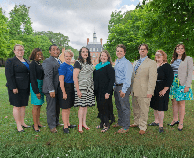 A group of people is standing together outdoors on a grassy area with trees and a building in the background.