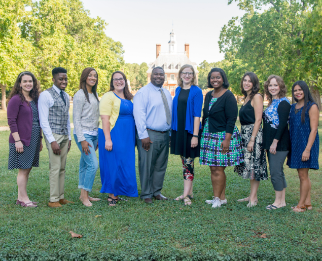 A group of people is standing on a grassy field with trees and a building in the background.