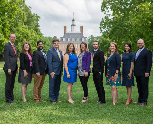 A group of people stands on a lawn in front of a historic brick building with a cupola, surrounded by lush green trees.