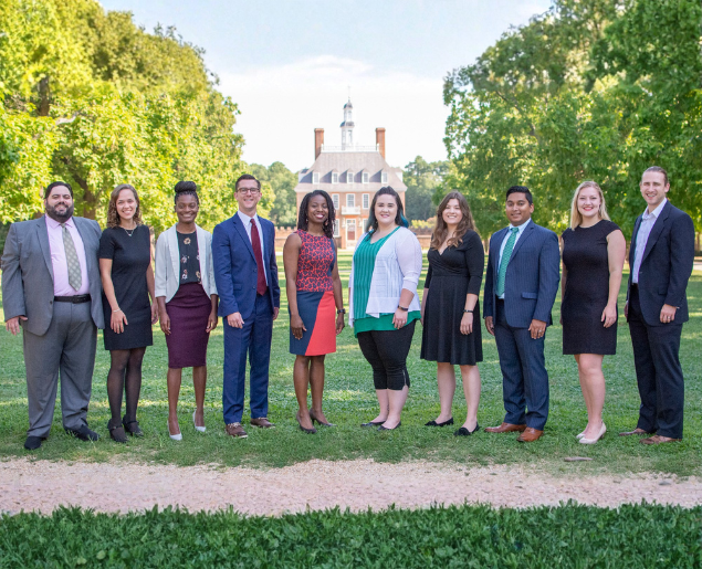 A diverse group of people dressed in business attire stands on a grassy path with a historic building and vibrant trees in the background.