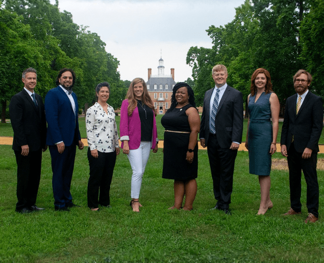 A group of people stands on a grassy pathway, flanked by tall trees and a historic building in the background.