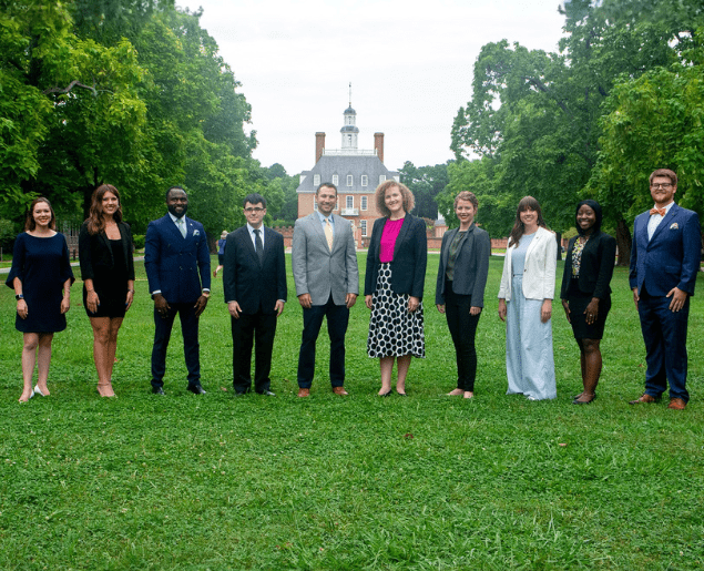 A group of people dressed in formal attire stands on a grassy area with a historic building and trees in the background.