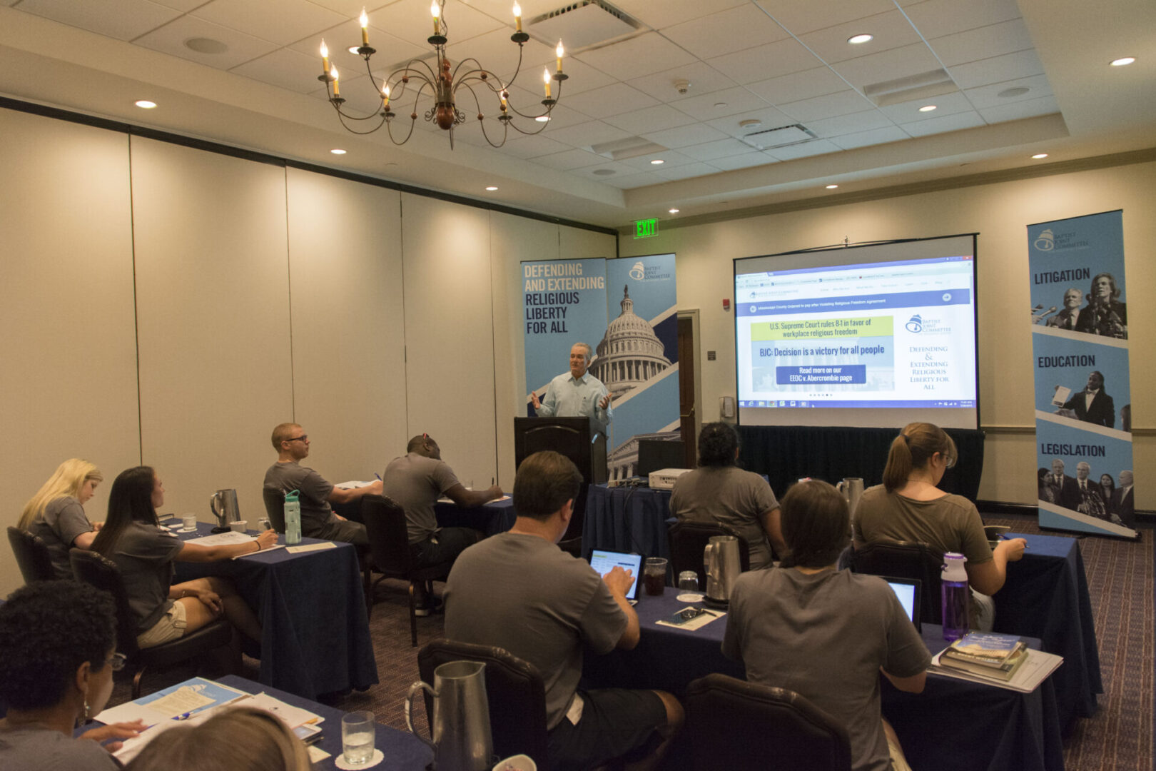 A person is giving a presentation in a conference room with attendees seated at tables.
