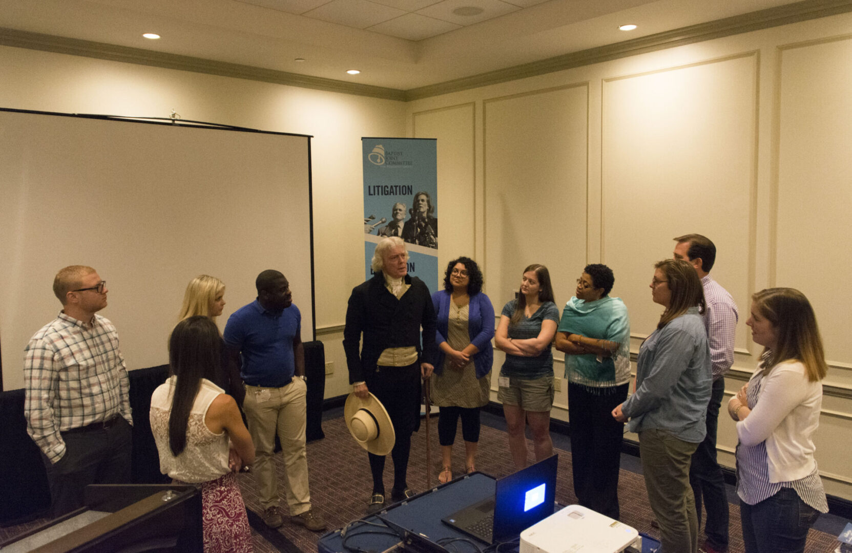 A group of people is gathered in a conference room, listening to a person in a cowboy hat speaking.