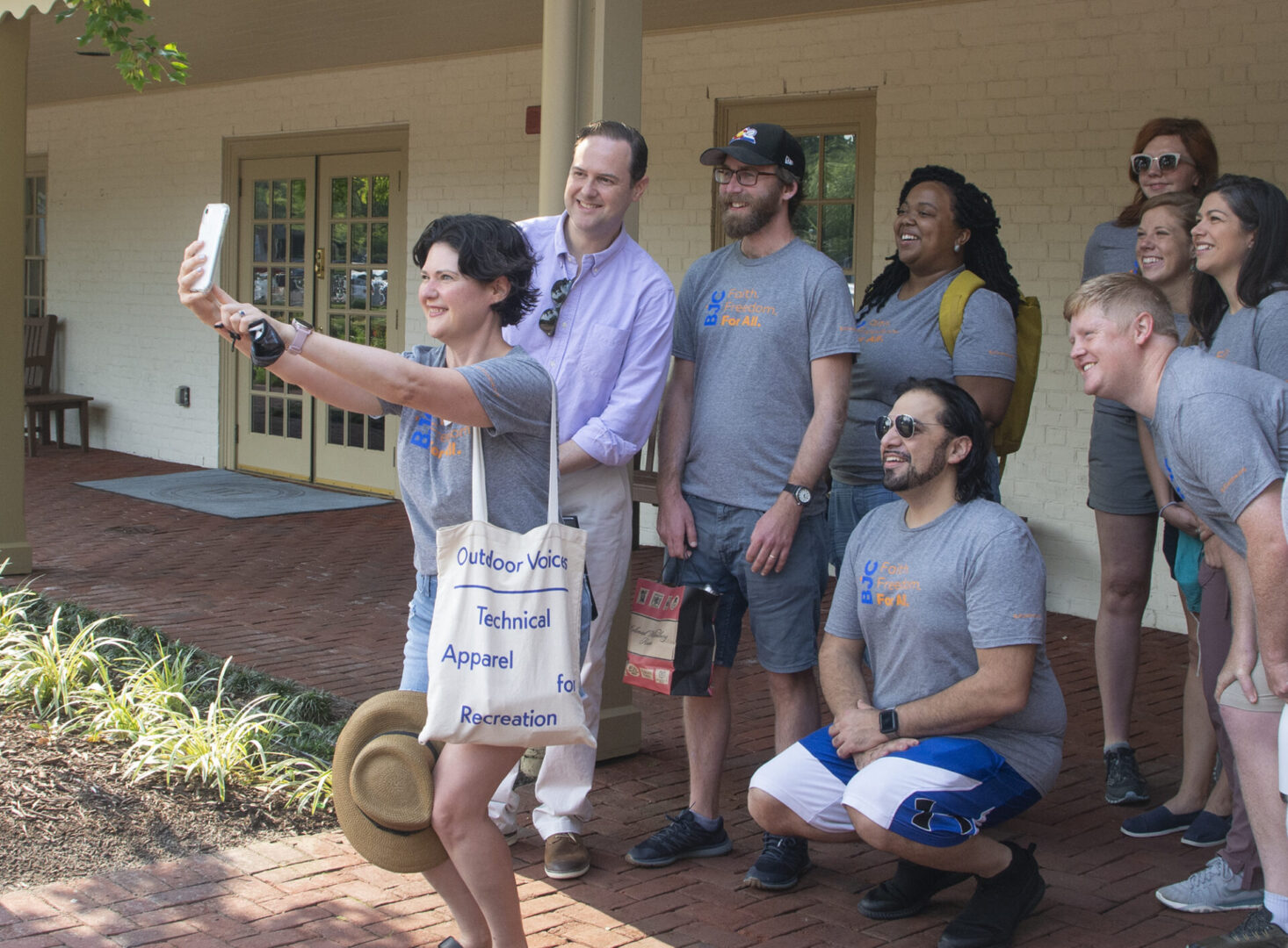A group of people in casual attire pose for a selfie on a brick pathway outside a building.