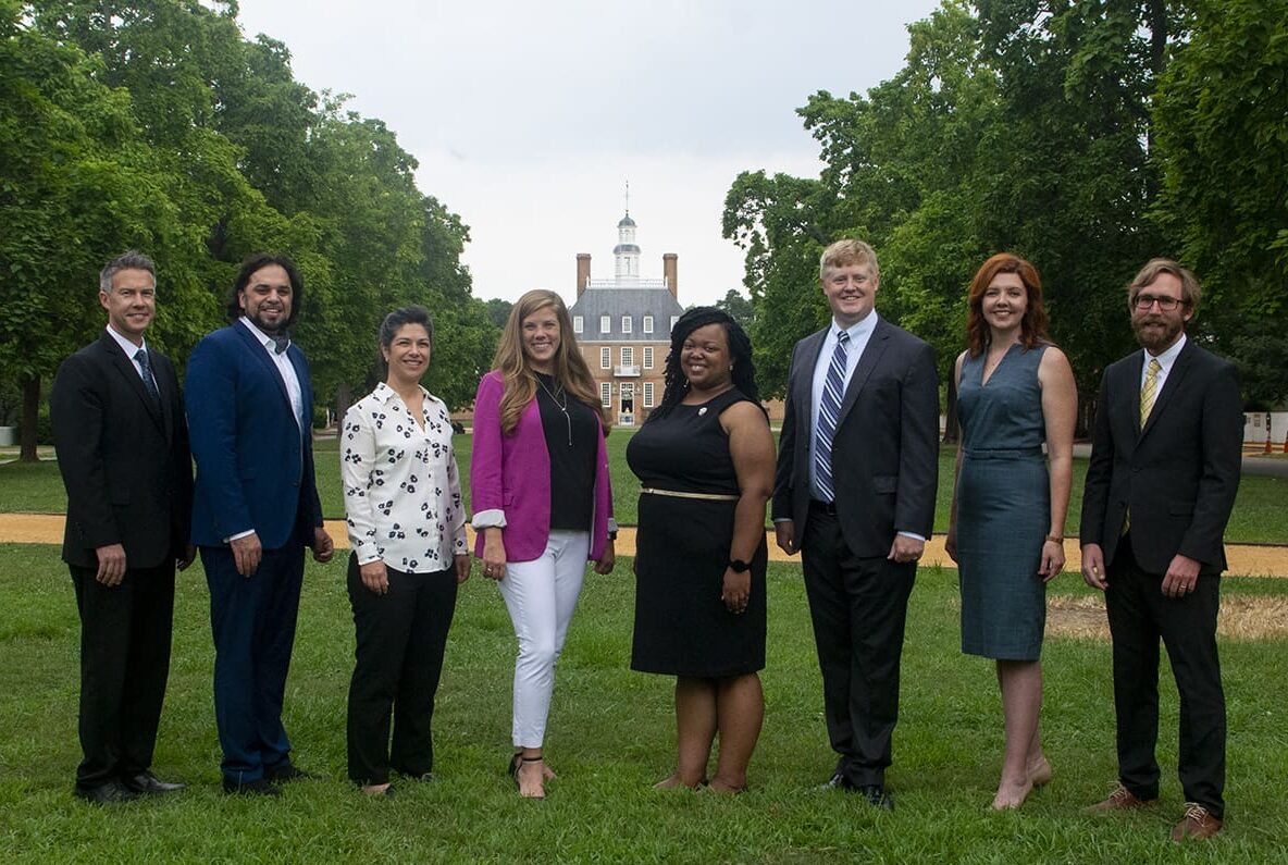 A group of people, including men and women in formal attire, stand on a grassy area with a historical building in the background.