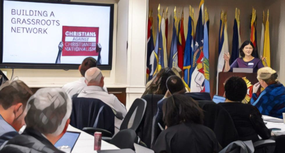 A group of people are seated in a room with various flags and a screen displaying a presentation.