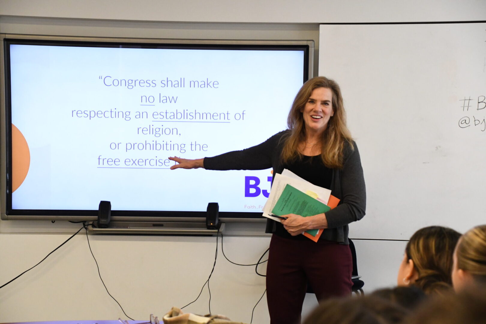 A woman stands in front of a screen giving a presentation and smiling, holding papers.