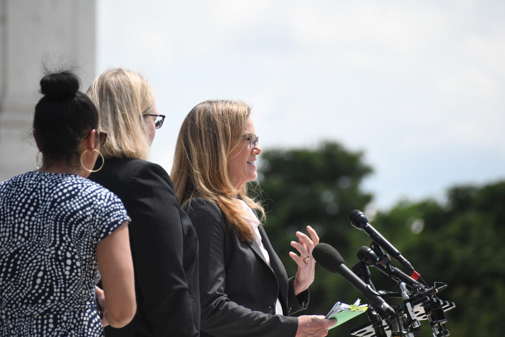A person is speaking at a podium with microphones outdoors, accompanied by others.
