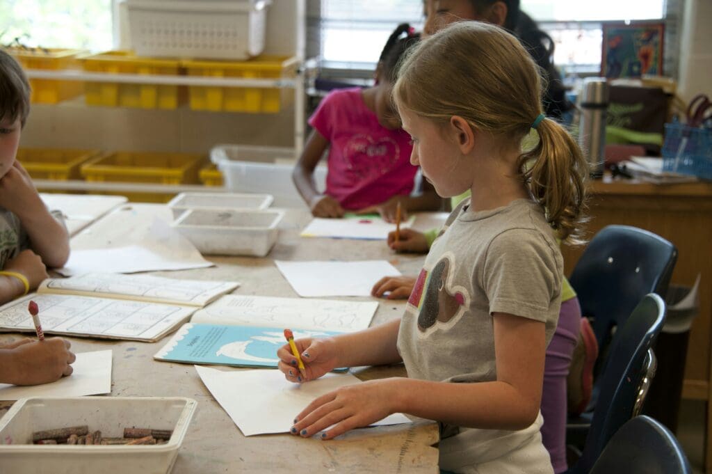 A child is sitting at a table, drawing or writing on paper with a pencil in a classroom setting.