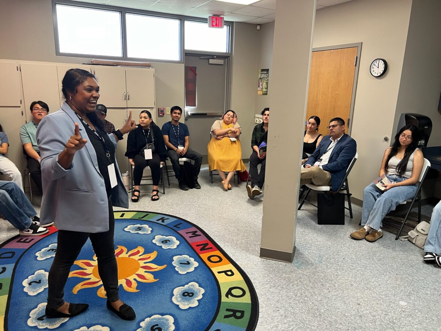A person is speaking to a group seated in a room with walls adorned with educational materials and a colorful alphabet rug on the floor.