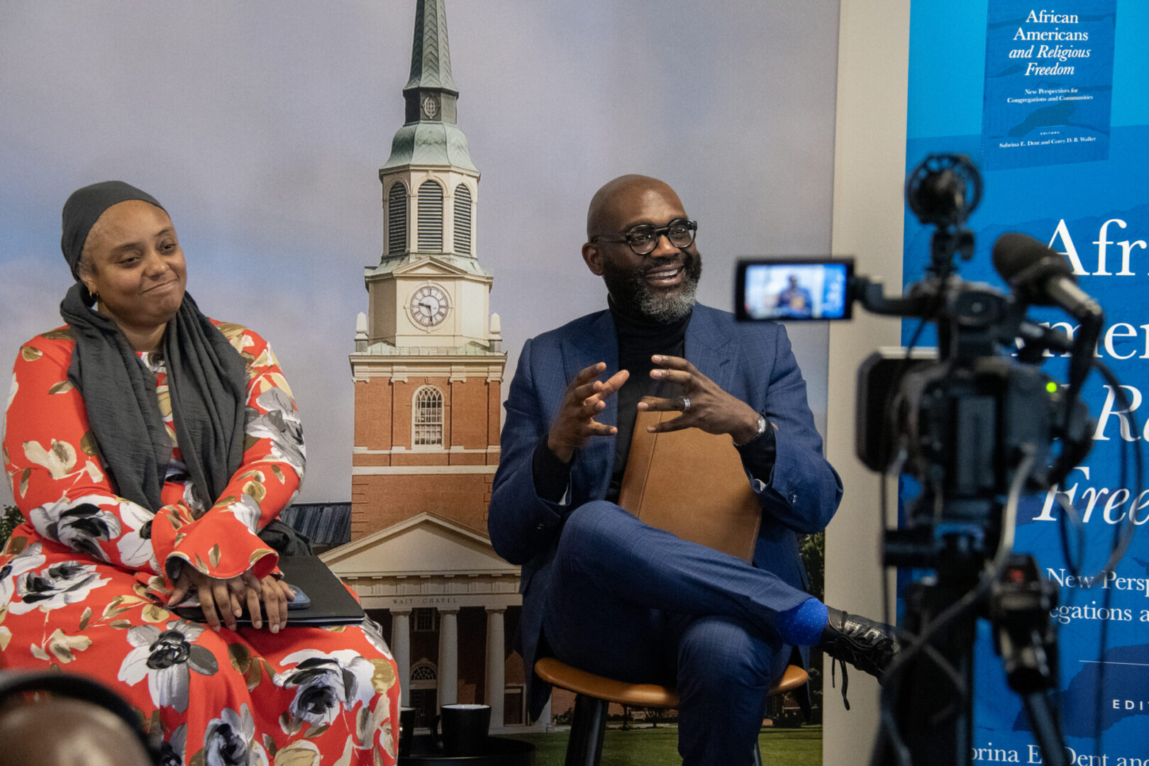 Two individuals are being filmed in a studio setting with a backdrop featuring a building with a tall steeple.
