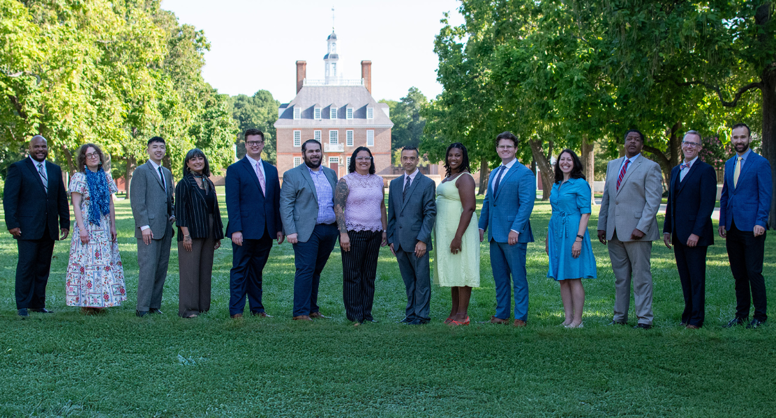 A group of people is standing together on a grassy area, with a historic building in the background and trees nearby.