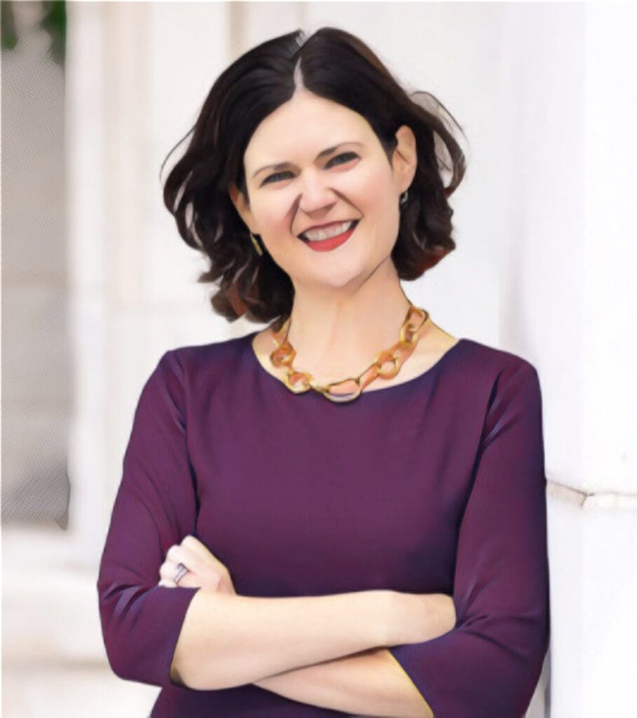 A person with short dark hair is smiling, wearing a purple top and a necklace, standing with arms crossed.