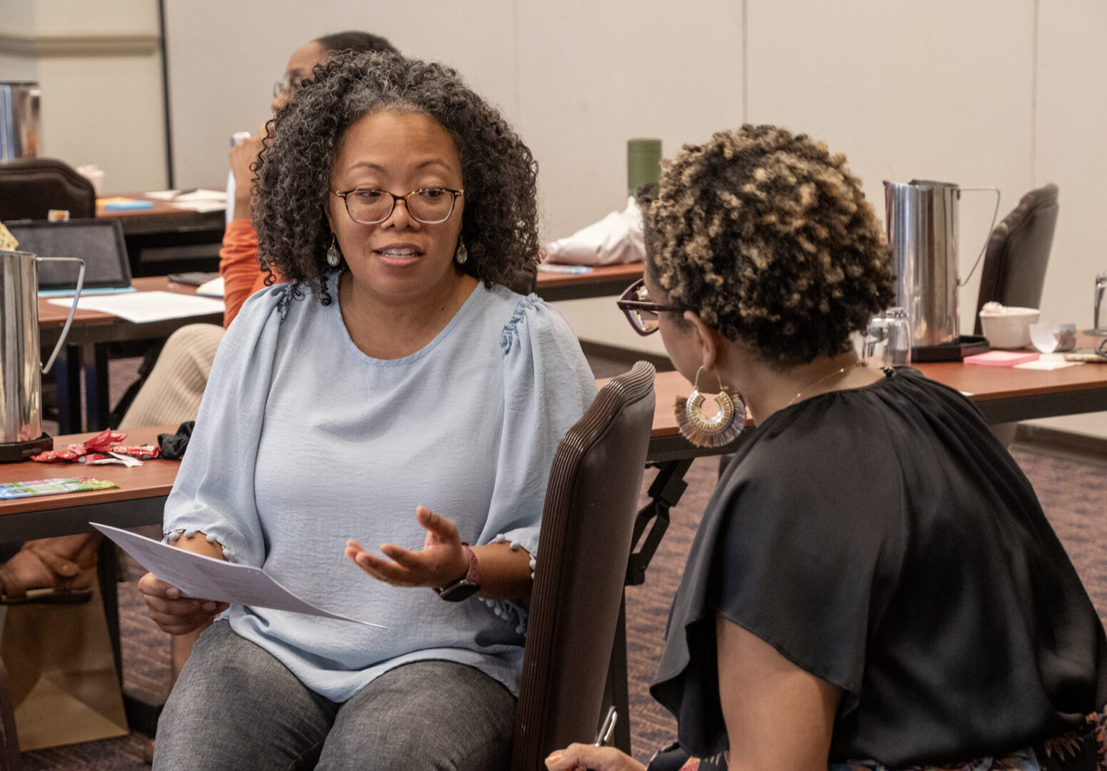 Two women are engaged in a conversation, with one holding papers.