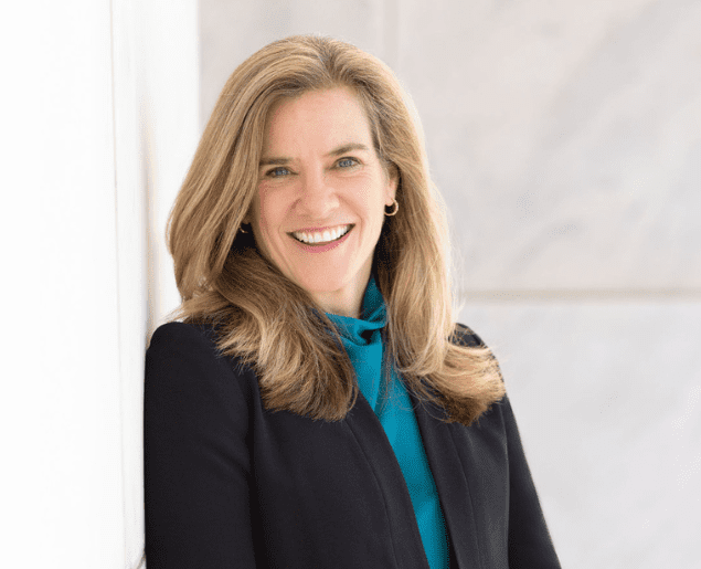 A person with long, light brown hair is smiling and wearing a dark blazer over a teal top.
