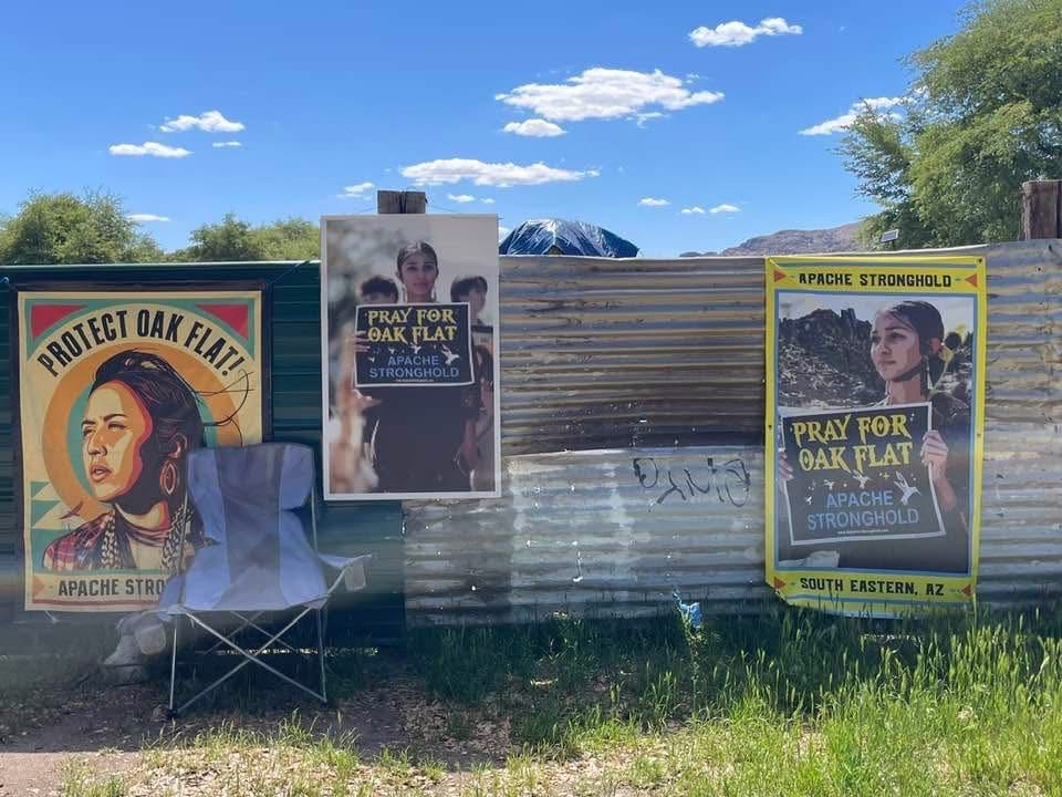 A corrugated metal fence is adorned with several posters and a folded chair in front, surrounded by greenery under a blue sky.